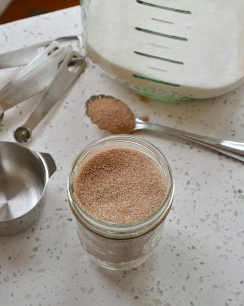 An overhead view of a mason jar and spoon full of cinnamon sugar with a measuring cup, measuring spoons, and sugar canister in the background.