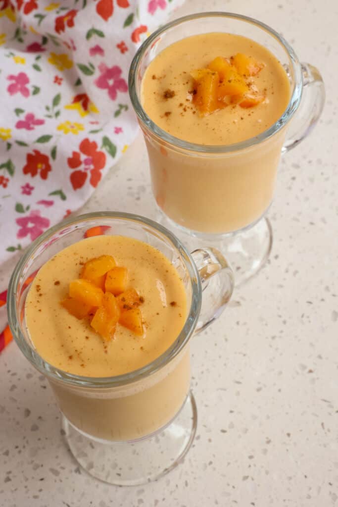 Two glasses of peach smoothie garnished with chopped peaches and a dash of cinnamon. 