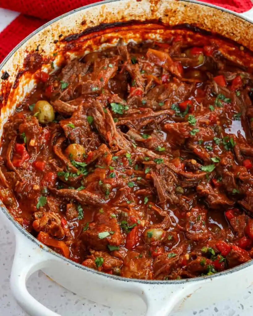 A Dutch oven full of ropa vieja with tender, slow-cooked shredded beef, onions, bell pepper, olives, and capers in a rich tomato-based sauce that is loaded with flavor. 