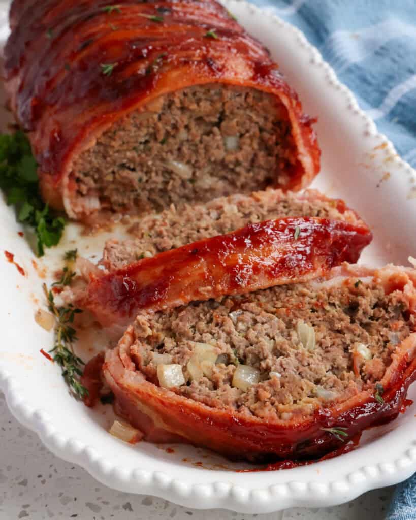 Sliced bacon wrapped meatloaf on a white serving platter. 