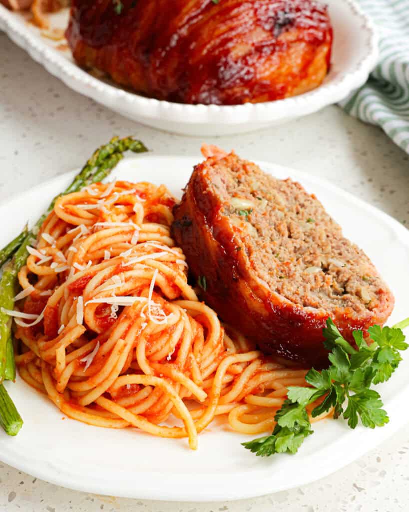 Bacon wrapped meatloaf on a serving plate with meatless spaghetti and oven roasted asparagus. 