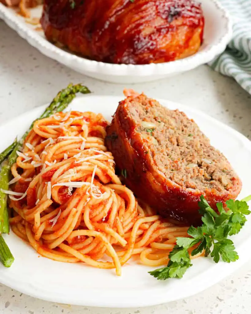 Bacon wrapped meatloaf on a serving plate with meatless spaghetti and oven roasted asparagus. 