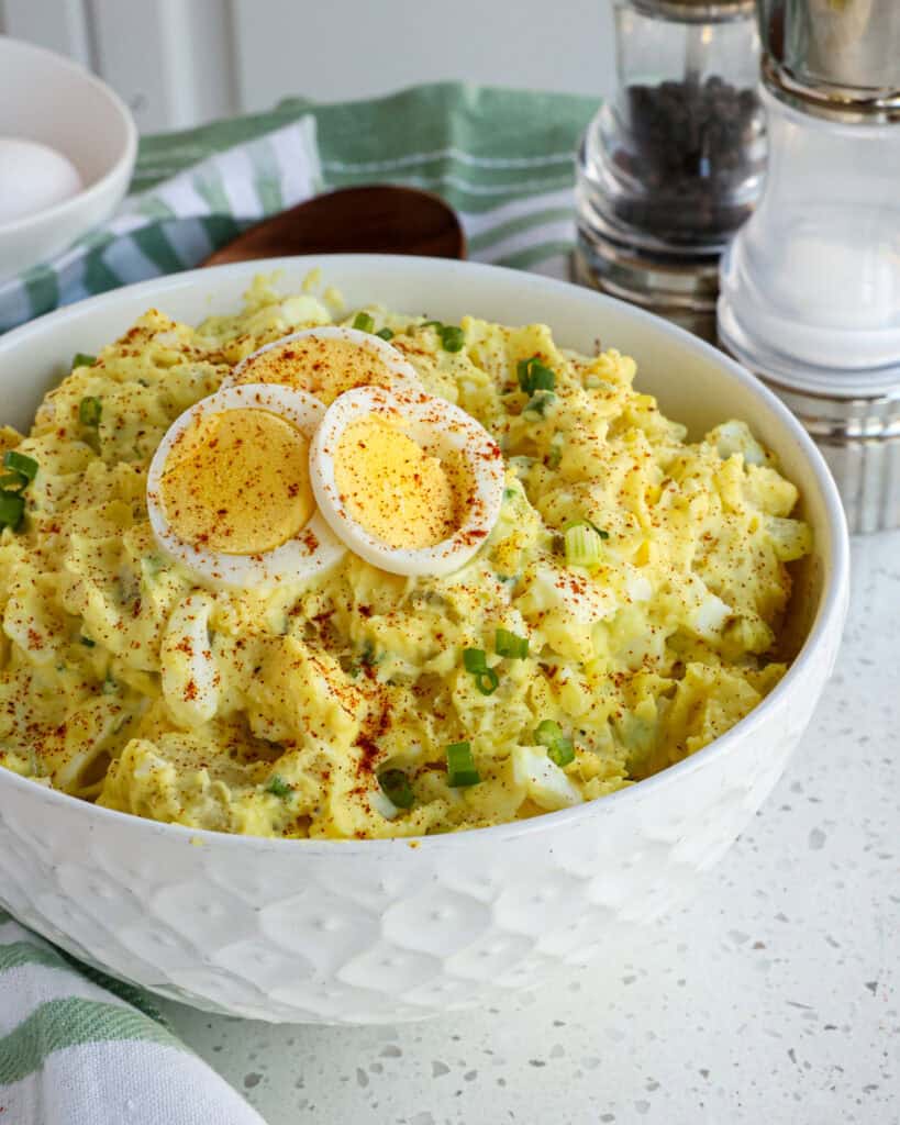 A serving bowl full of southern potato salad with salt and pepper, hard cooked eggs, and a wooden spoon the background. 