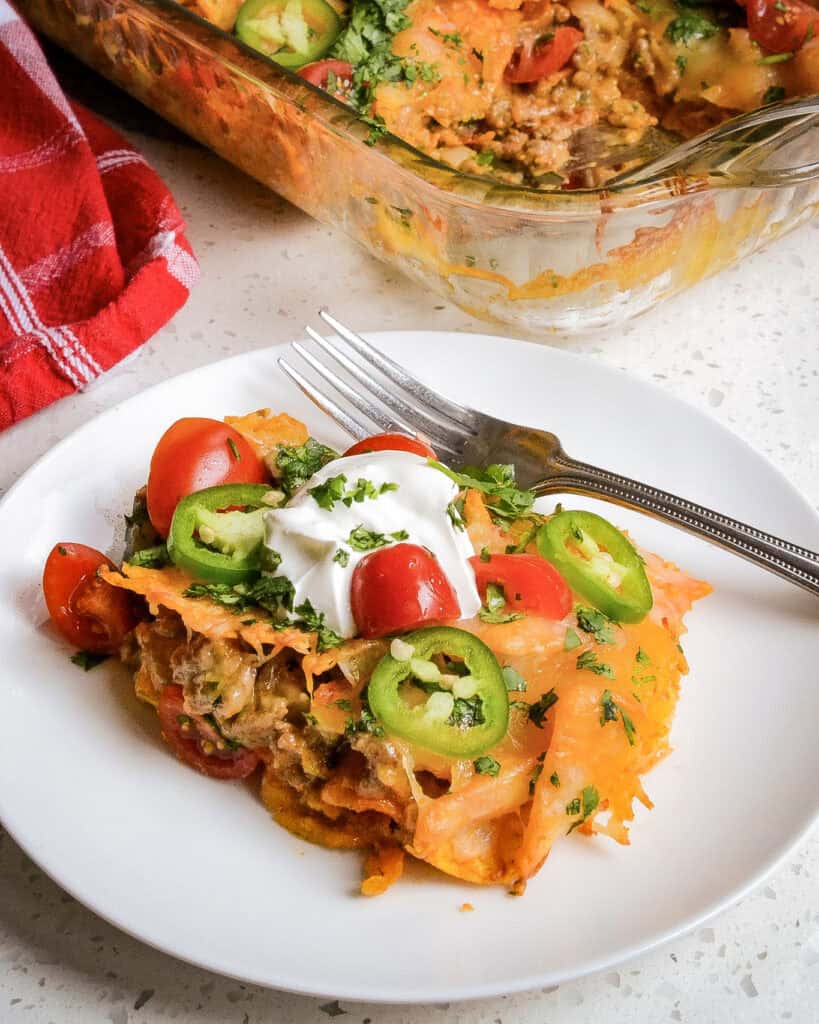 Doritos casserole on a single serving plate with a fork in front of a casserole dish filled with Doritos casserole.