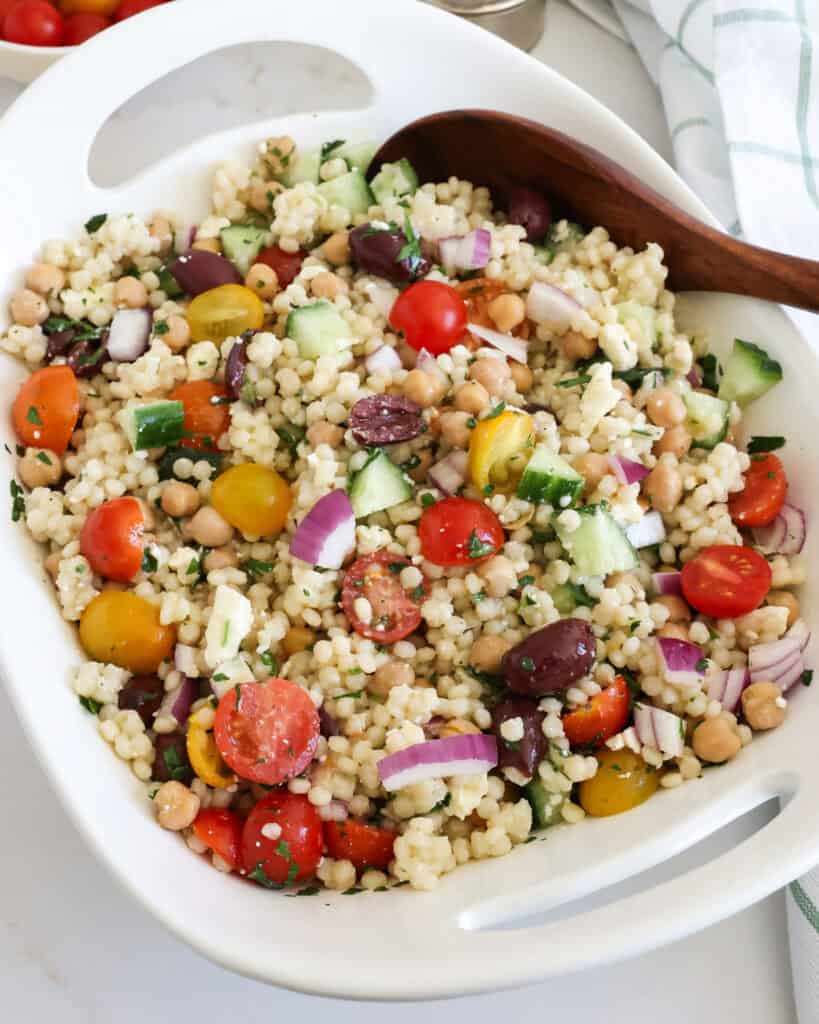Couscous salad in a bowl with a wooden spoon with tomaotes, lemon, and salt and pepper shaker in the background. 