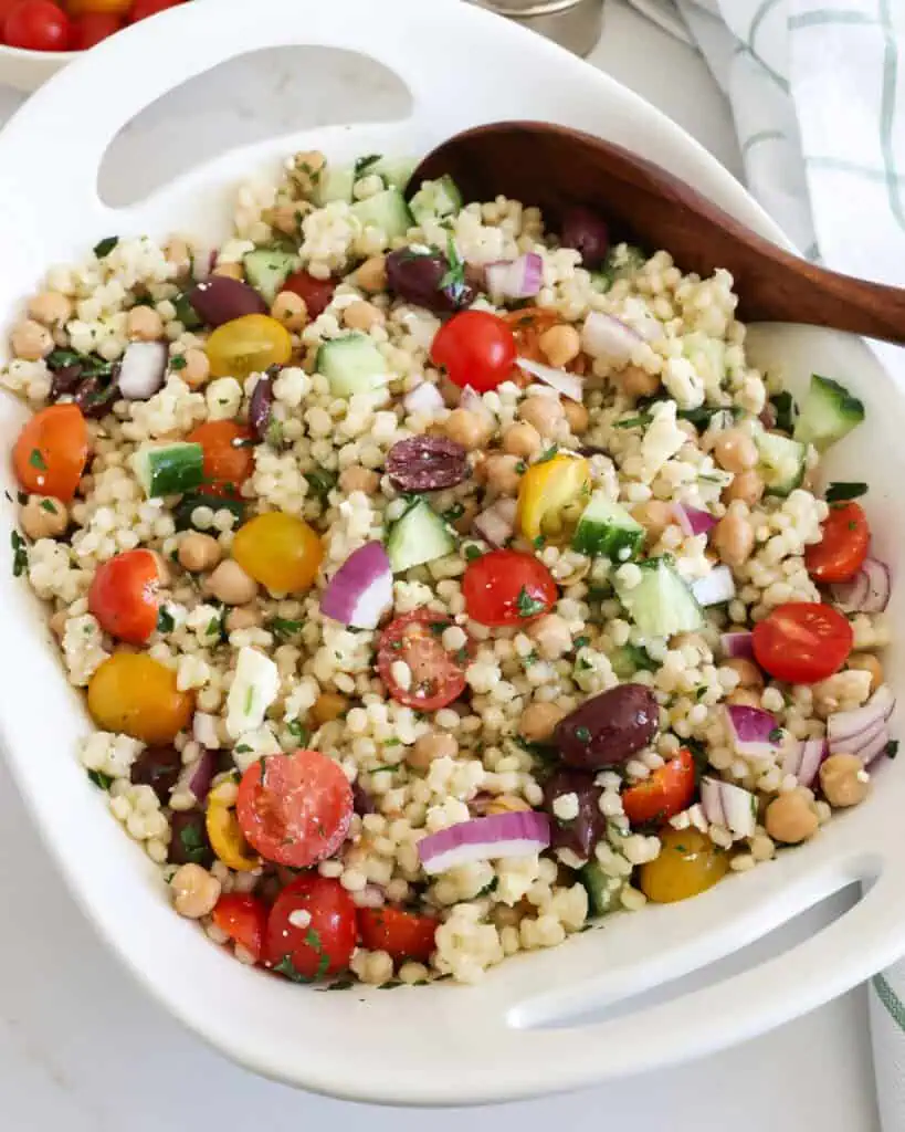 Couscous salad in a bowl with a wooden spoon with tomaotes, lemon, and salt and pepper shaker in the background. 