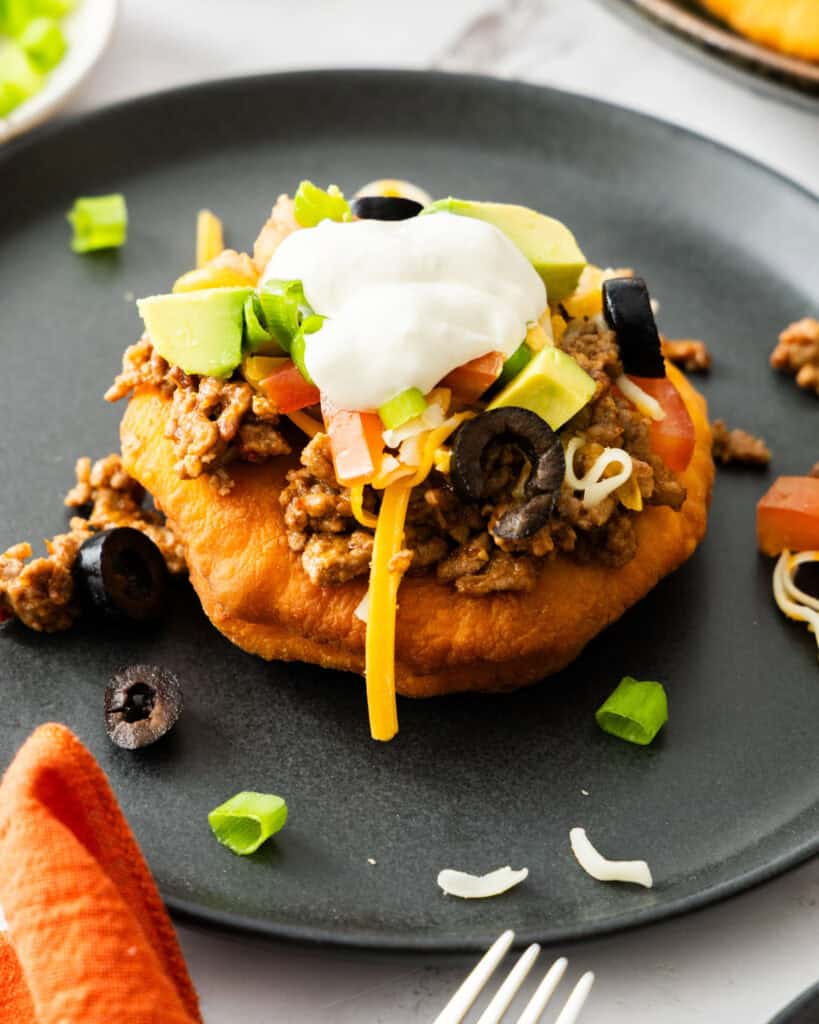 An overhead view of a Navajo taco topped with cheddar cheese, tomatoe, avocado, and sour cream on a single serving black plate.