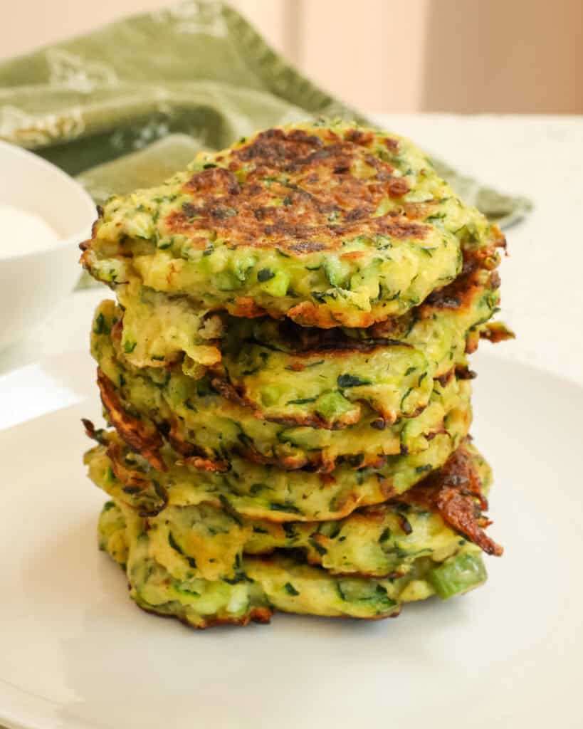 Zucchini Fritters stacked on a white plate with a green towel behind it.