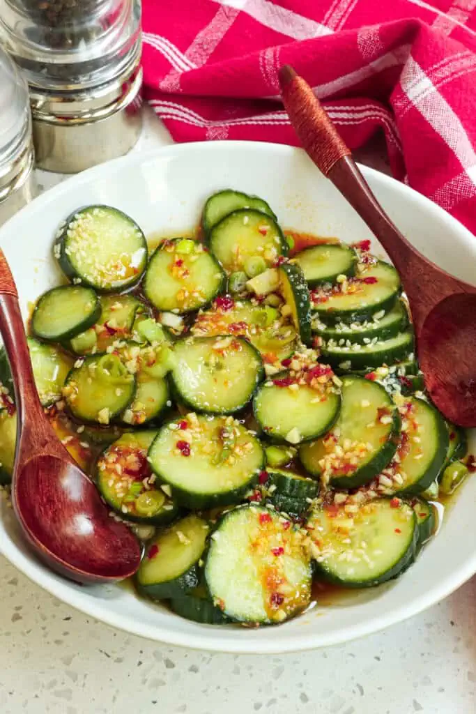 Zesty Asian cucumber salad in a bowl with two wooden spoons and a salt and pepper shaker on the side of the bowl. 

