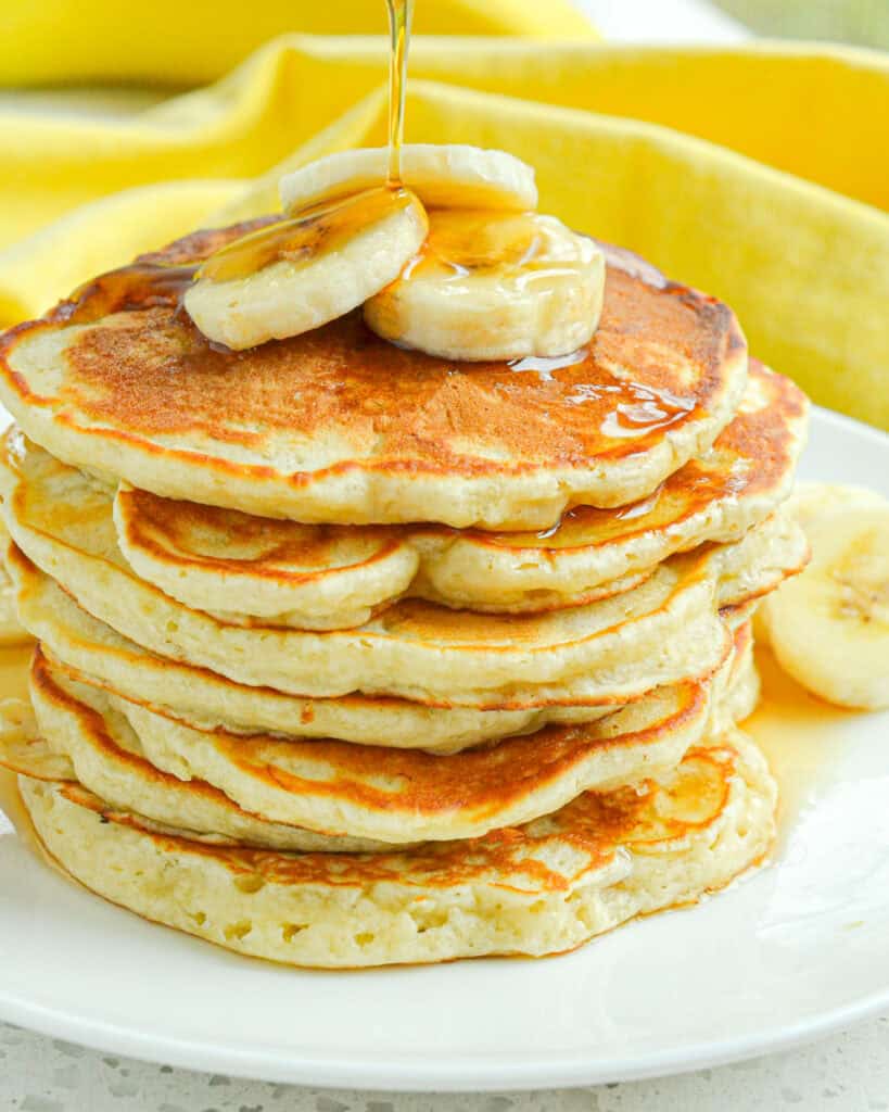 A close up view of banana panakes with syrup and slices of banana on a white plate.