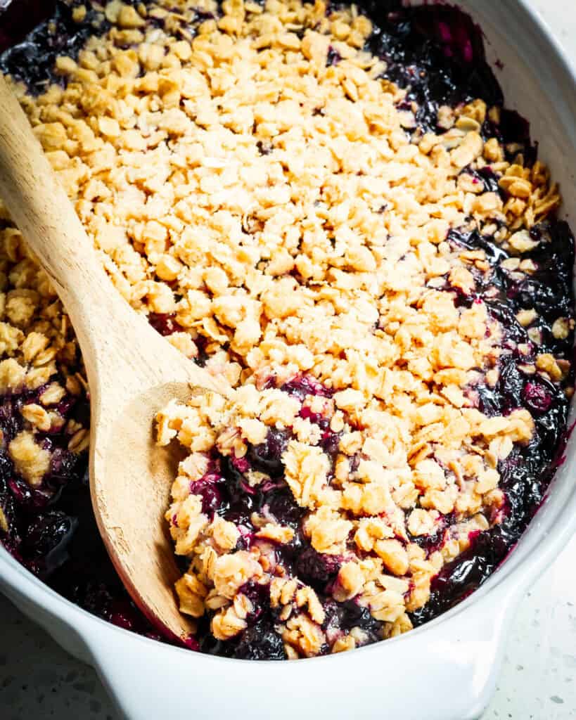 Blueberry crisp in an oblong white casserole dish with a wooden spoon. 