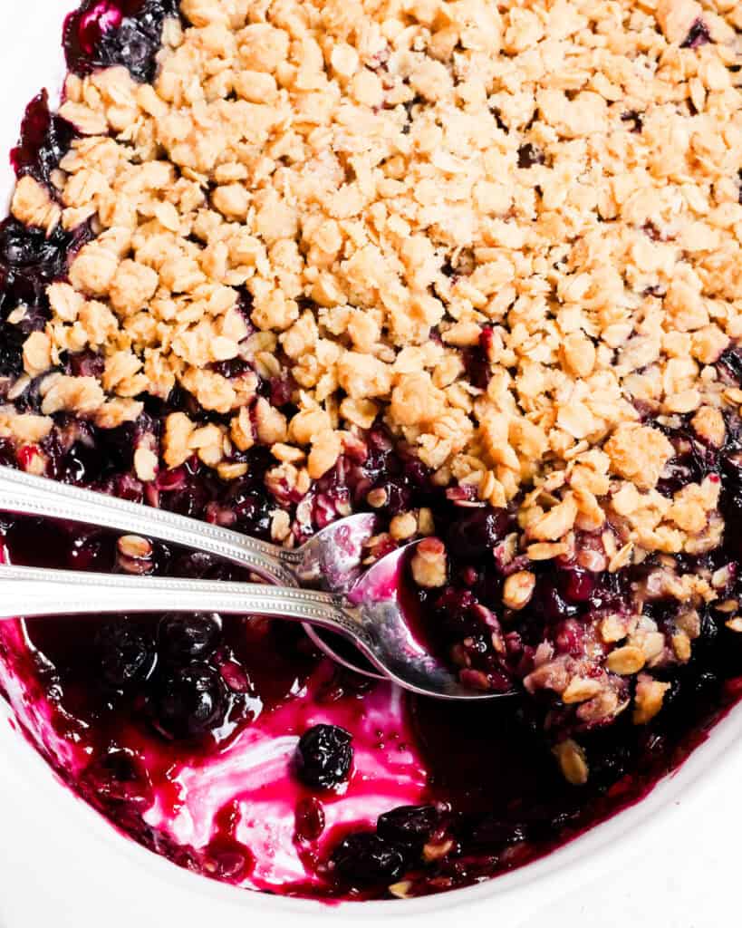 An oblong casserole dish full of blueberry crisp with 2 spoons in it. 
