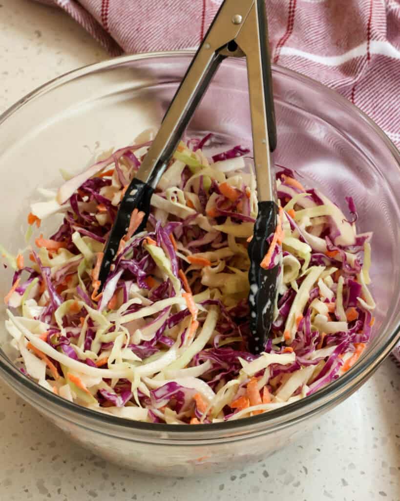 A clear bowl with a pair of kitchen tongs in it full of tri-color coleslaw mix and coleslaw dressing. 