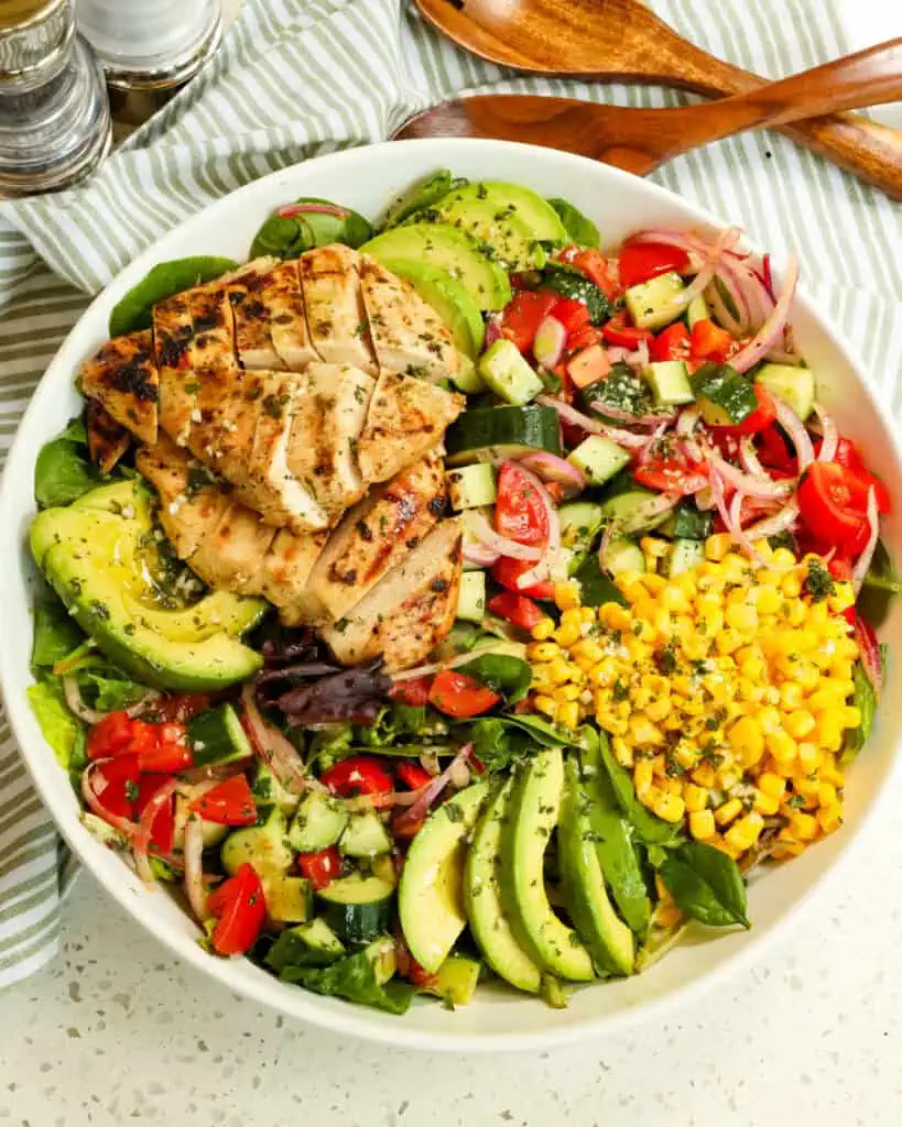 An overhead view of grilled chicken salad with corn tomatoes, avocado, cucumber and red onion.