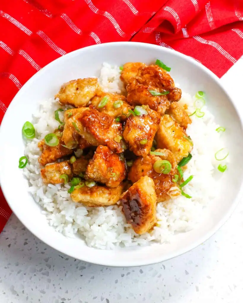 An overhead view of honey garlic chicken and green onions over white rice in a white bowl. 
