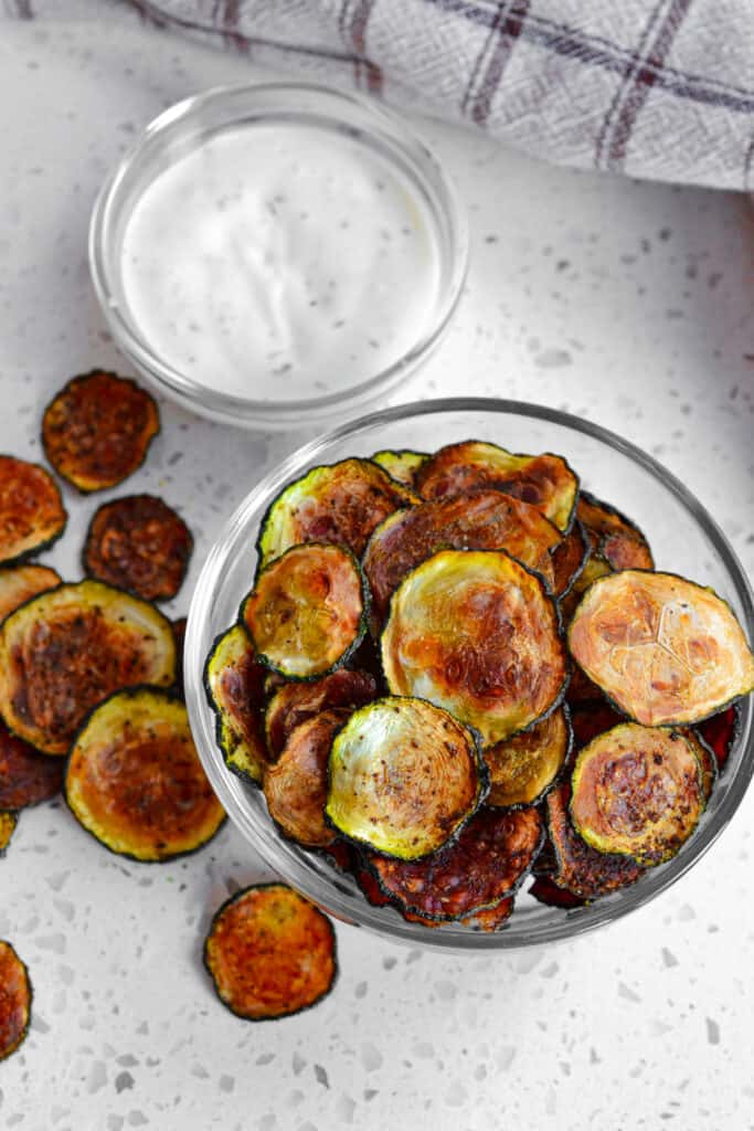 A glass bowl full of zucchini chips next to a smaller glass bowl full of homemade ranch dressing. 