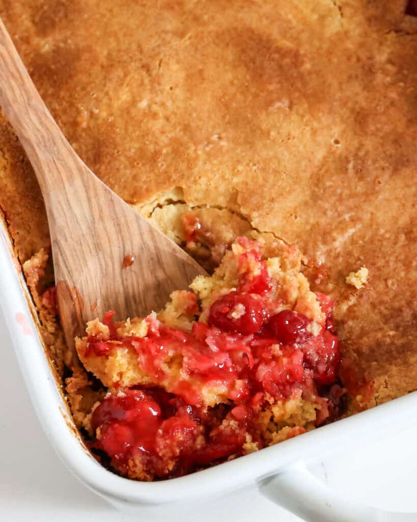A baking dish and wooden spoon full of cherry dump cake. 