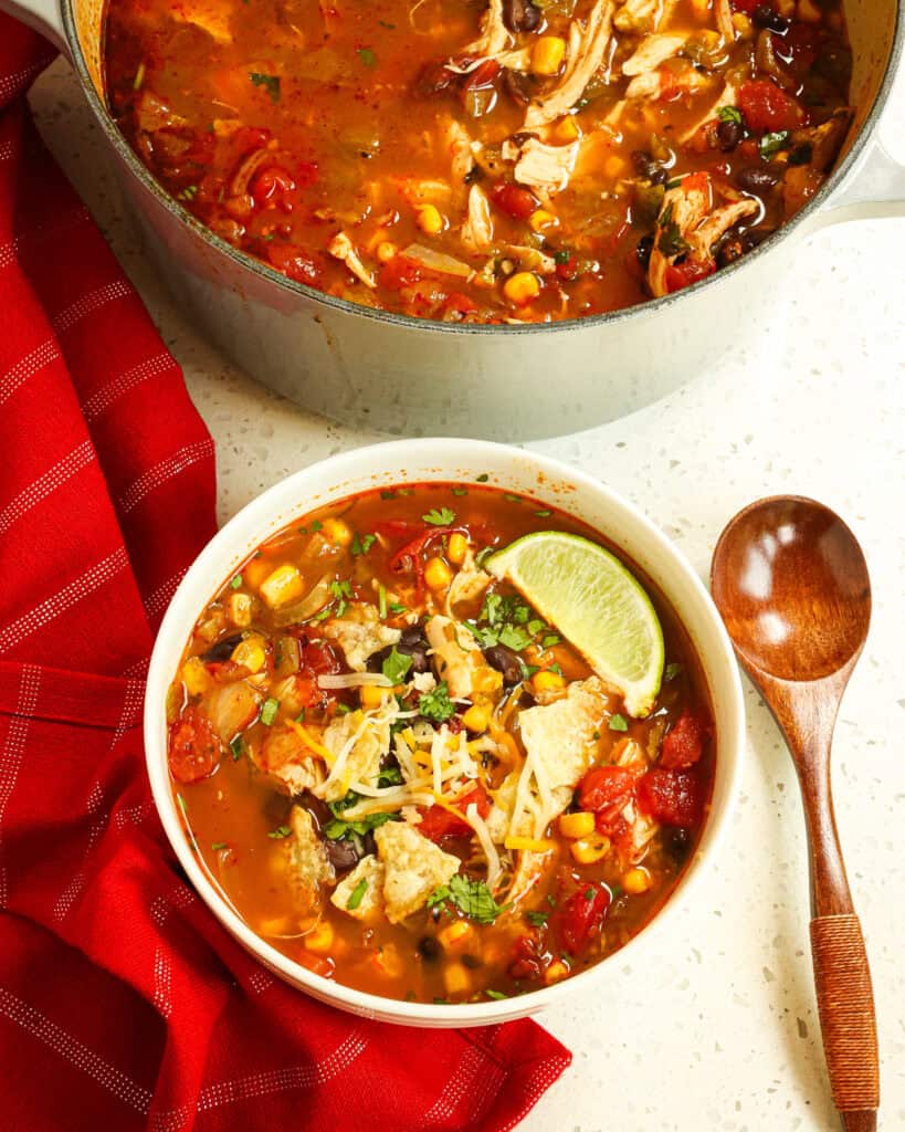 An overhead view of a single serving bowl full of chicken taco soup. with a wooden spoon and a Dutch oven fuill of the soup in the background. 