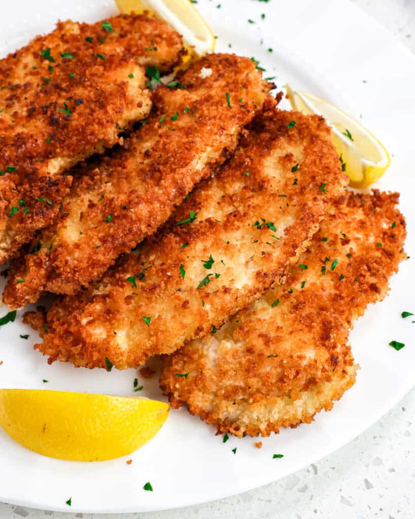 Multiple pieces of chicken schnitzel on a plate with lemon wedges. 
