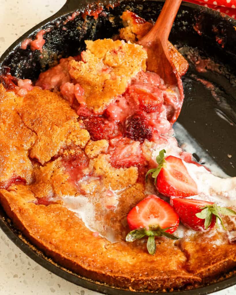 Close up view of strawberry cobbler in a cast iron skillet with fresh halved strawberries on top.
