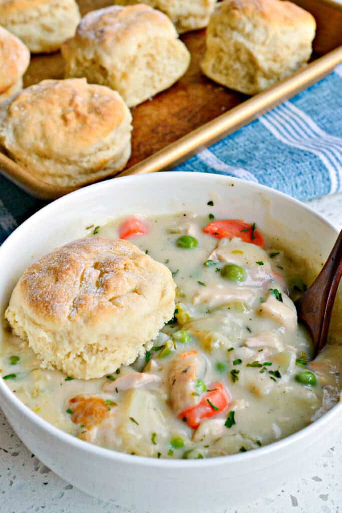 A single serving bowl full of chicken pot pie soup with a biscuit on the top and baking try full of biscuits in the background. 