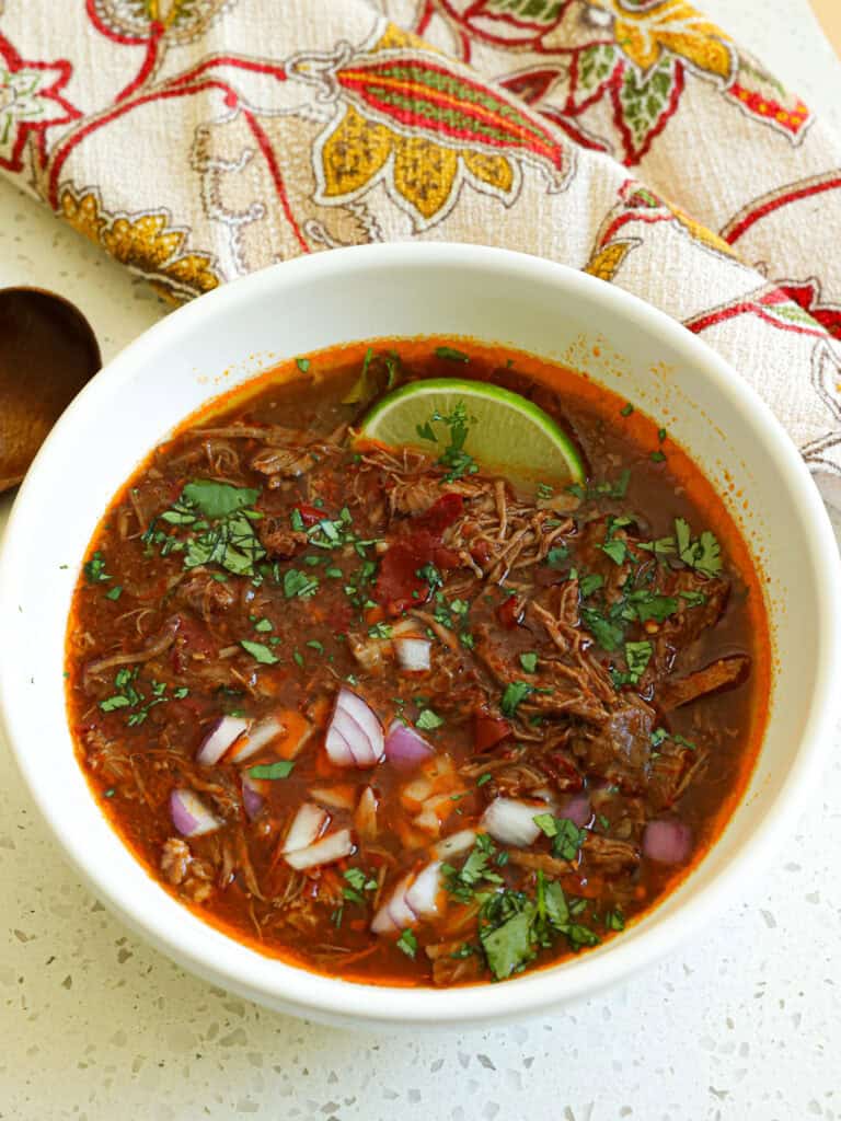 Beef birria in a white serving bowl with limes, chopped cilantro, and chopped red onions. 