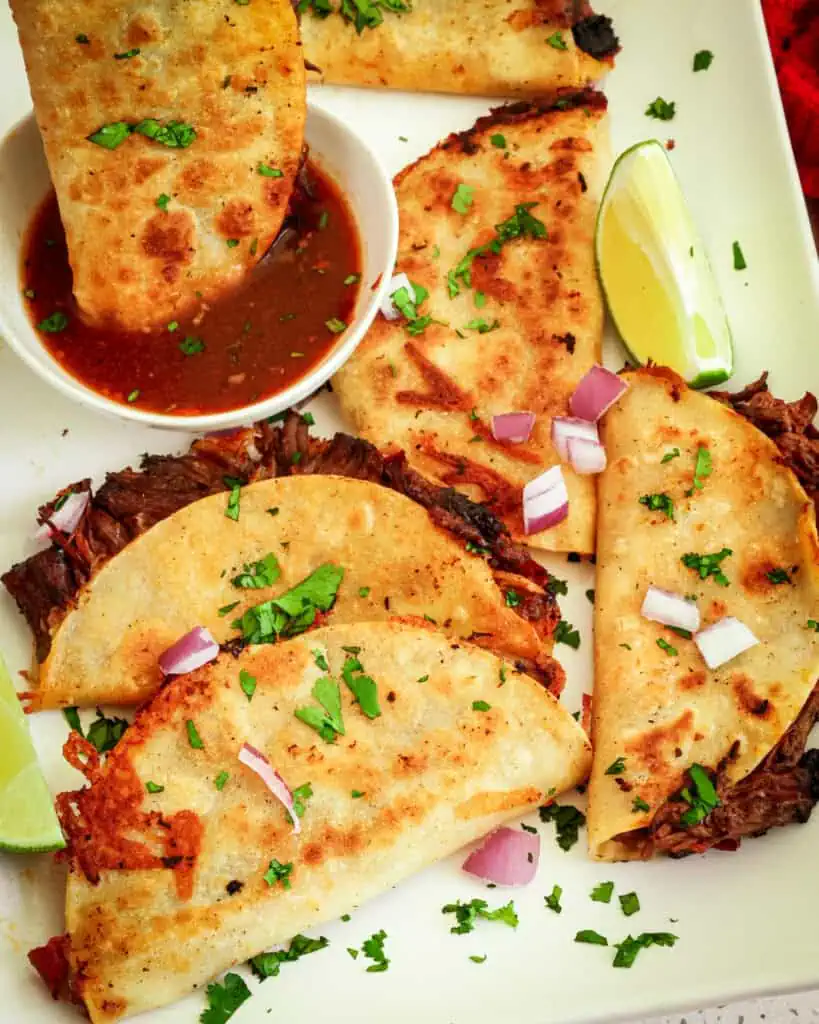 A platter full of birria beef tacos with limes, red onions, chopped cilantro and dipping sauce.