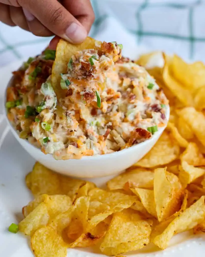 A bowl of million dollar dip with a potato chip dipped in it on a plate full of potato chips. 