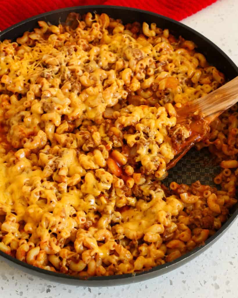 A large black skillet and a wooden spoon full of beefaroni smothered in melted cheddar. 
