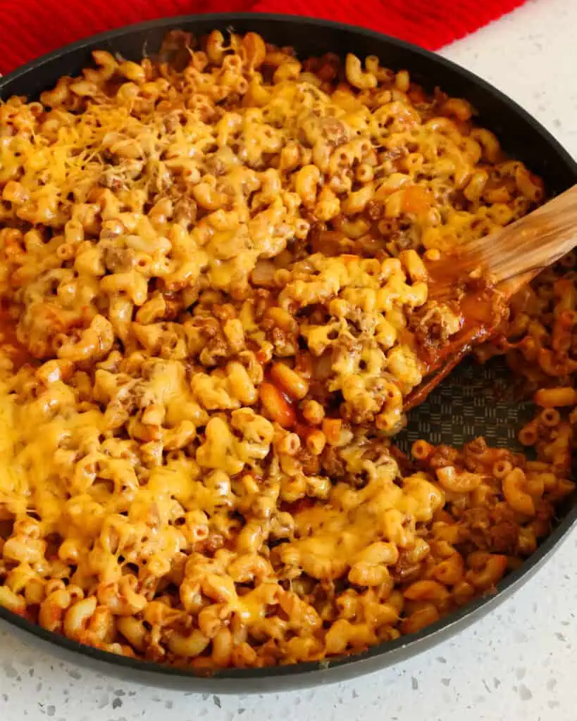 A large black skillet and a wooden spoon full of beefaroni smothered in melted cheddar. 