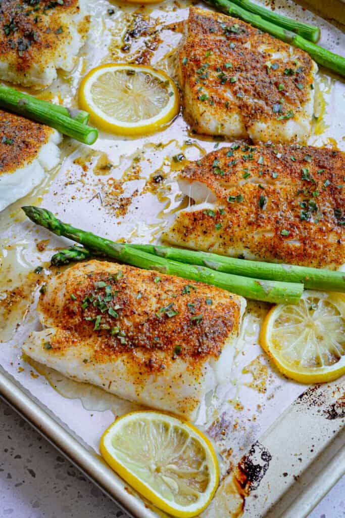 Cod filets on a baking sheet with lemon slices and asparagus. 