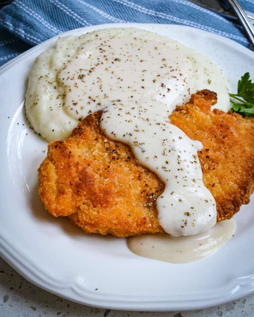 Country gravy over fried chicken breast and mashed potatoes on a single serving white plate. 