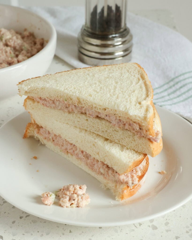 A double stacked deviled ham sandwich on a single serving white plate with a bowl of deviled ham salad in the background. 