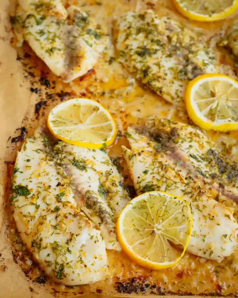 Fresh baked lemon dip tilapia with lemon slices and herbs on a parchment covered baking sheet. 