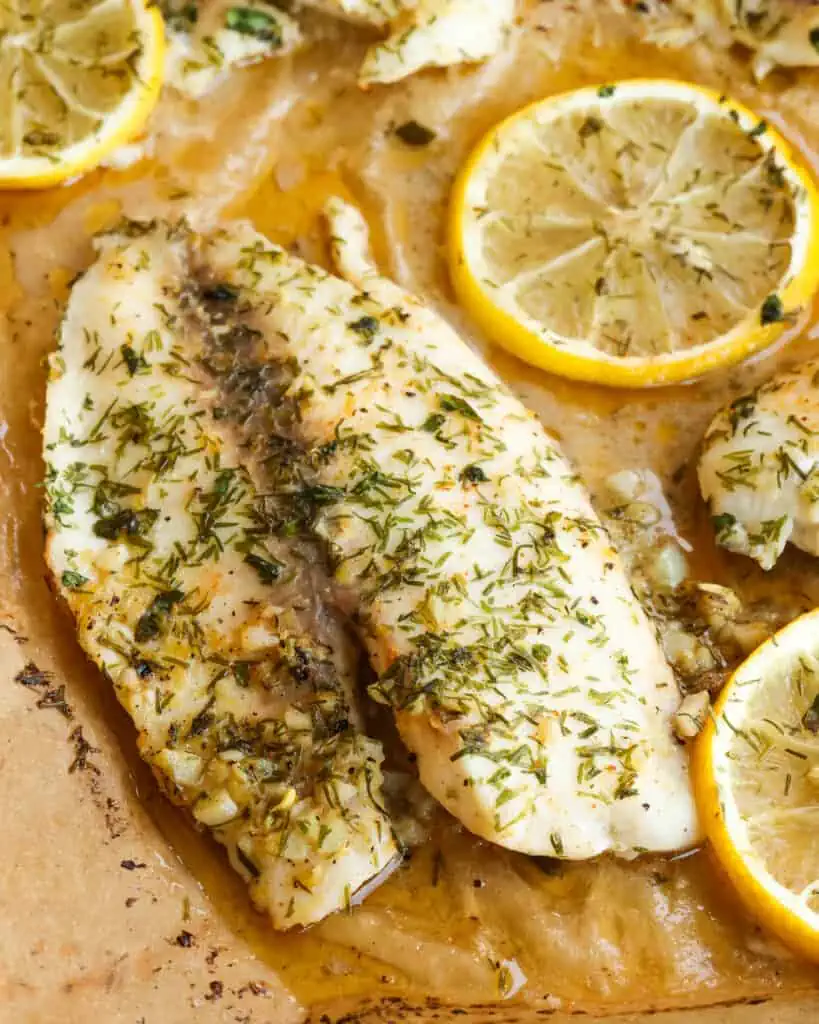 A close up view of baked tilapia with fresh herbs and lemons on parchment paper. 