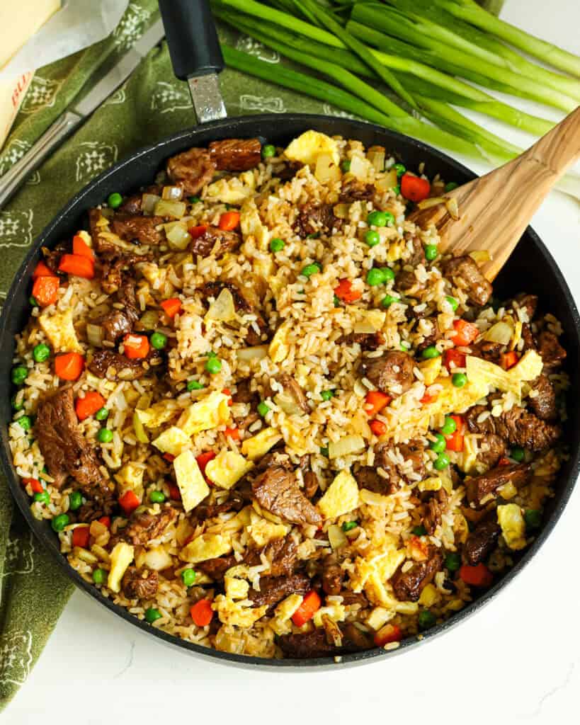 An overhead view of beef fried rice in a large skillet with a wooden spatula. 