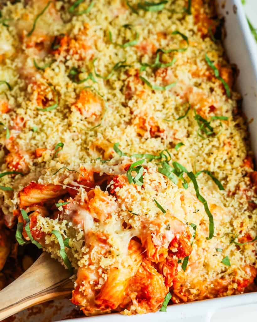 A white casserole dish and a wooden spoon full of chicken parmesan casserole. 