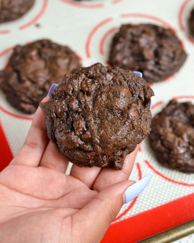 A hand holding up a double chocolate chip cookie over a baking sheet with more double chocolate chip cookies. 