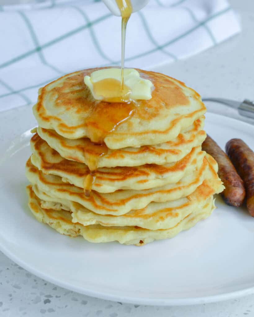 A stack of oatmeal pancakes with butter and maple syrup and two sausage links on a single serving plate.