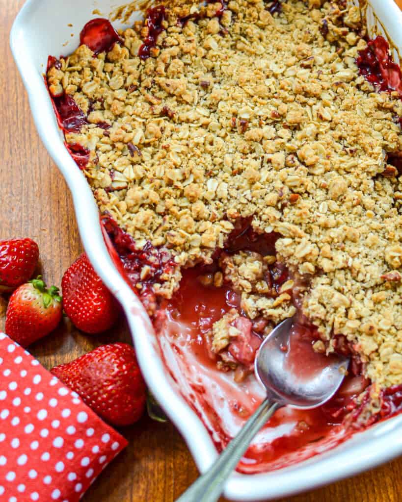 Overhead shot of Strawberry Rhubarb Crisp with a spoon in the white dish and strawberries to the left.