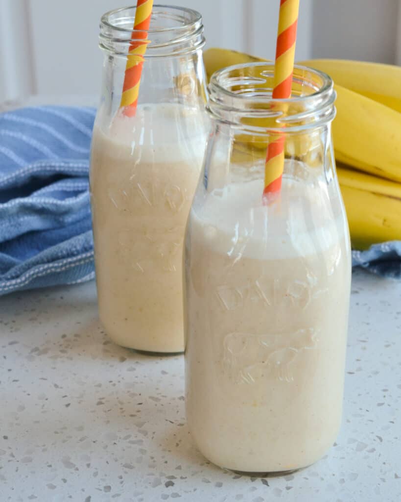 Two single serving milk glasses full of banana milk and orange and yellow straws and bananas in the background.