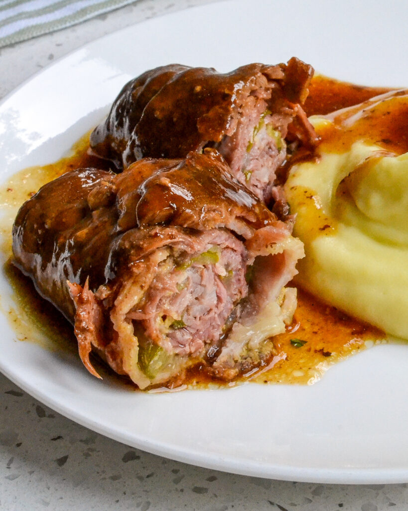 German Beef Rouladen cut in half to show the inside with mashed potatoes off to the side and gravy on top.