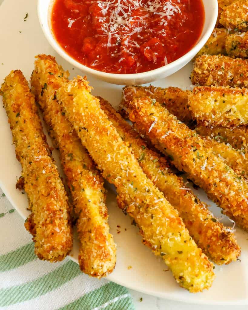 A close up view of freshly fried zucchini on a plate with a bowl of marinara. 