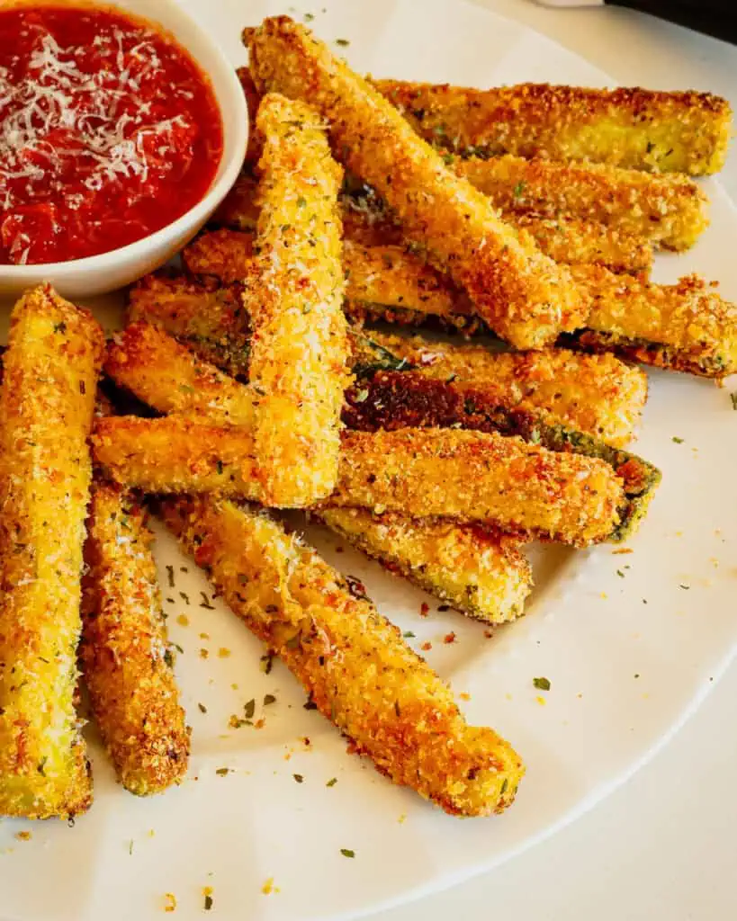 Fried zucchini sticks on a whtie plate with a small bowl of marinara.