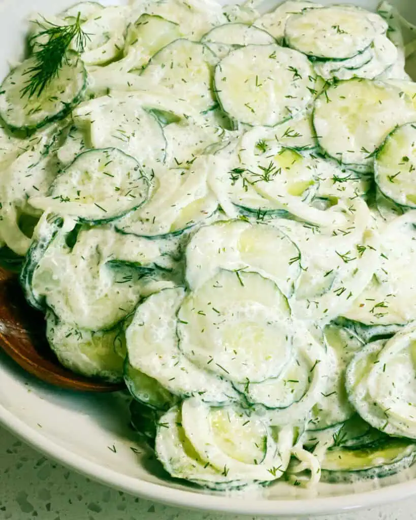 A close up view of German cucumber salad in a whiter serving bowl with a wooden spoon. 
