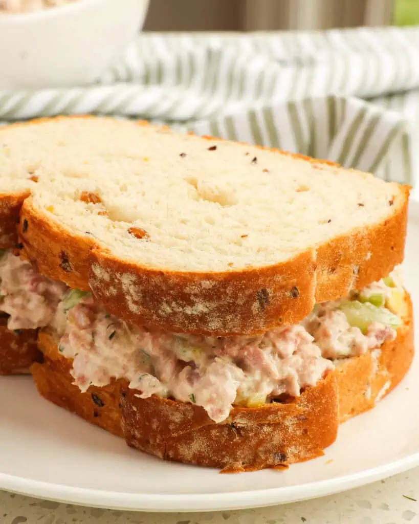Ham salad on white bread on a single sserving plate with a green and white striped towel in the background. 