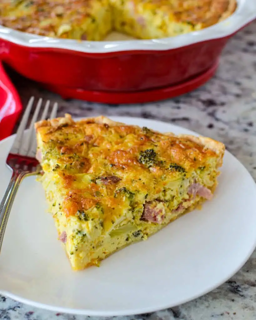 A slice of ham and cheese quiche on a single serving plate with a fork. 