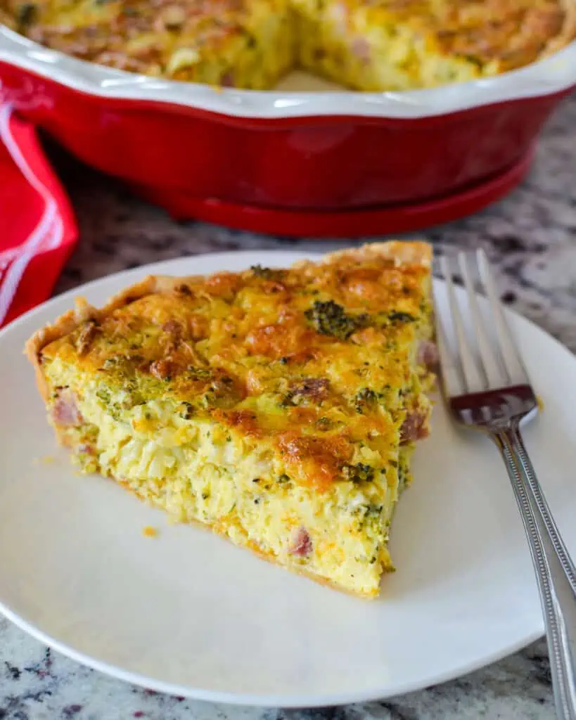 A slice of ham and cheese quiche on a serving plate with a fork and the rest of the quiche in the background. 
