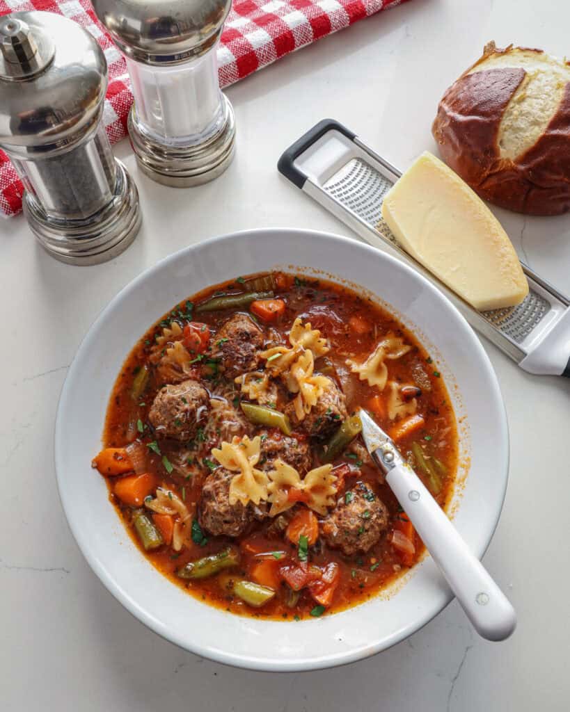 An overhead view of a single serving white soup bowl full of meatball soup with a salt and pepper shaker, block of Parmesan cheese and grater, and a loaf of pretzel bread in the background. 