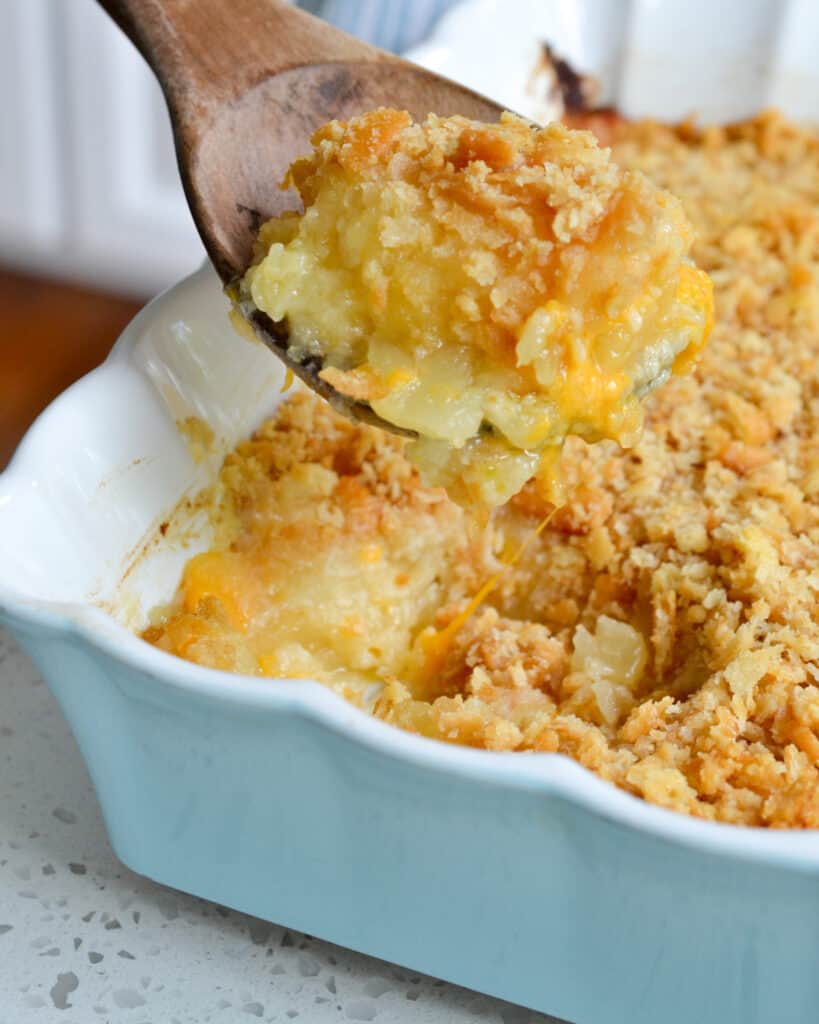 A wooden spoon full of pinapple casserole over a casserole dish full of pineapple casserole. 